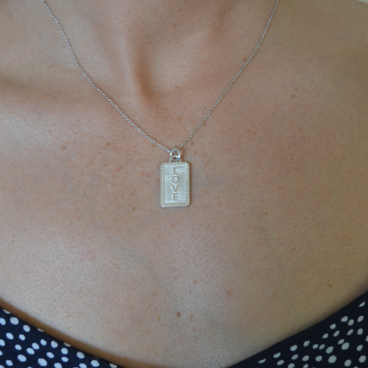 Our handcrafted sterling silver charm with raised lettering. This side says AMOR. It is shown with a 16" necklace on a model.